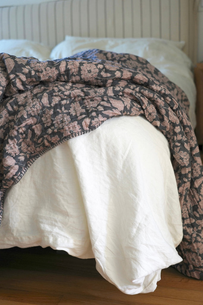 Anthracite Flower Sherpa Cottage blanket with charcoal and lilac floral design, casually draped over the side of a bed to show its cozy texture and plush Sherpa lining 